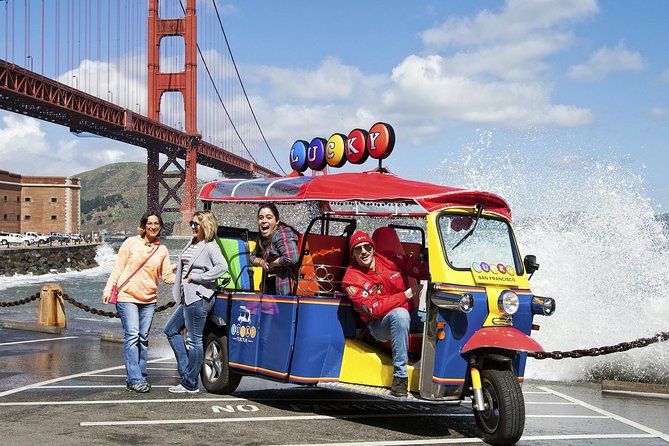 A group of three women smiling and laughing in a colorful blue and red electric Lucky Tuk Tuk parked at Fort Point with the Golden Gate Bridge in the background.