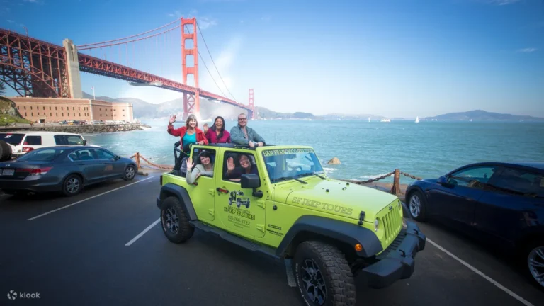 A group of five smiling tourists posing in a bright green open-top San Francisco Jeep Tours vehicle at Fort Point, with the Golden Gate Bridge and San Francisco Bay in the background.